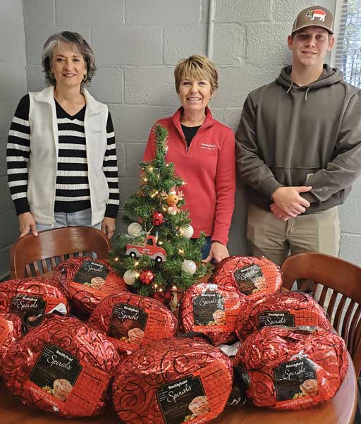 Three people standing behind a table with a small Christmas tree and several wrapped hams