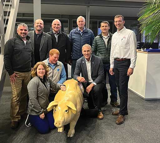 6 adults standing and 3 kneeling to pose around a yellow statue of a pig