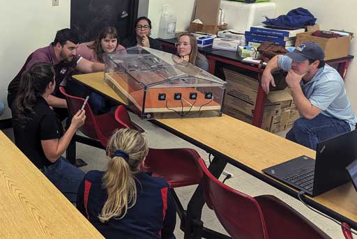 Several people around a table looking at a model of a barn made from transparent plastic panels